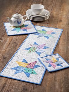 two quilted placemats on a wooden table with teapot and saucer