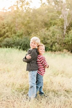 two young boys hugging each other while standing in tall grass with trees in the background