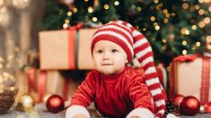 a baby wearing a red and white striped hat sitting in front of a christmas tree