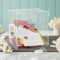 a clear box filled with lots of envelopes on top of a table next to flowers