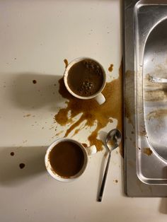 two cups of hot chocolate sit on a dirty counter next to a metal tray with spoons