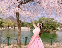 a woman in a pink and white dress standing under a cherry blossom tree next to a lake