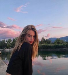 a woman standing next to a body of water with mountains in the background at sunset