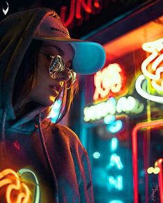 a woman standing in front of a neon sign at night wearing sunglasses and a hat