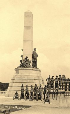 black and white photograph of men sitting on steps in front of a monument with a man standing at the base