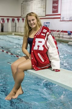 a woman sitting on the edge of a swimming pool wearing a varsity jacket and shorts