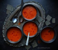 four bowls of red soup with spoons and crackers on a tray, top view