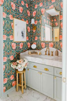 a bathroom with floral wallpaper and marble counter tops, gold faucet, vanity mirror, and stool