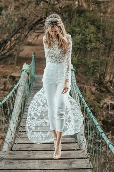 a woman is walking across a bridge wearing white pants and a lace top with flowers in her hair