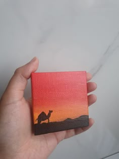 a hand holding up a small painting with a camel in the sunset on it's side