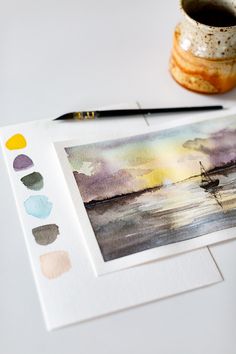 a watercolor painting on paper next to a cup and paintbrush, with the image of a sailboat in the distance