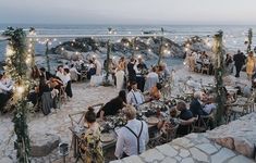 a group of people sitting at tables next to the ocean with string lights strung over them