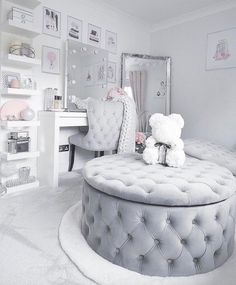 a white bedroom with a round ottoman and teddy bear