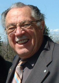 an older man wearing glasses and a suit smiling for the camera with trees in the background