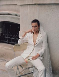 a woman sitting on top of a chair next to a wall wearing a white suit