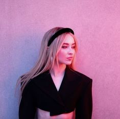 a woman with long blonde hair wearing a black jacket and headband standing in front of a pink wall