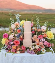 a table topped with a pink cake surrounded by lots of flowers and greenery next to a river