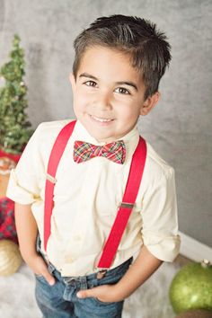 a young boy wearing a bow tie and suspenders standing in front of a christmas tree