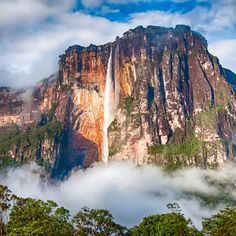 a tall waterfall towering over a forest filled with green trees and clouds in the sky