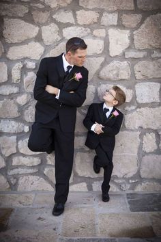 two young boys dressed in black suits and bow ties, standing against a stone wall