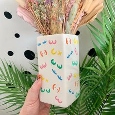 a hand holding up a vase with flowers in it and palm leaves on the side