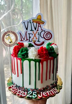 a hand holding a decorated birthday cake in front of a window with the words viva mexico on it