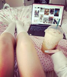 a woman is sitting in bed with her legs crossed and holding a drink next to her laptop