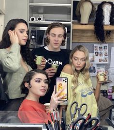 three women standing in front of a counter holding coffee cups and looking at their cell phones