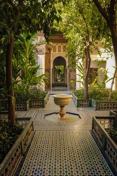 a fountain in the middle of a courtyard surrounded by trees and bushes, with an archway leading to it