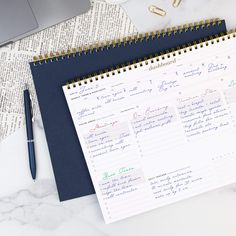 an open planner next to a laptop on a marble table with a notepad and pen