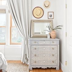 a white dresser sitting next to a window covered in curtains and pictures on the wall