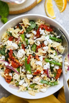 a white bowl filled with pasta salad and topped with feta cheese, red onions, green peppers, and lemon wedges