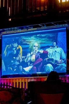 people sitting in chairs watching a movie on a large screen at an outdoor event with neon lights