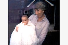 an older woman holding a baby wearing a white coat in front of a house with the door open