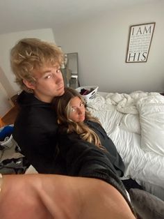 two people laying on a bed in a room with white sheets and pillows, one person has his arm around the other's head