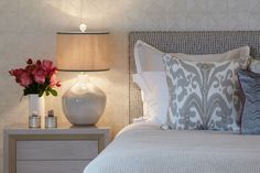 a white vase with pink flowers sitting on a nightstand next to a lamp and pillows