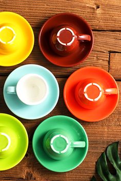 four colorful cups and saucers sitting on top of a wooden table next to a plant