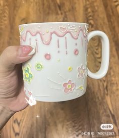 a hand holding a coffee mug with pink frosting and flowers on the outside, in front of a wooden table