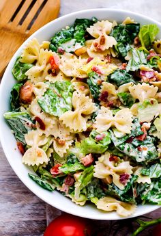 a white bowl filled with pasta salad next to tomatoes and lettuce on a wooden table