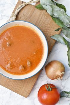 a bowl of tomato soup on a cutting board next to two tomatoes and an onion