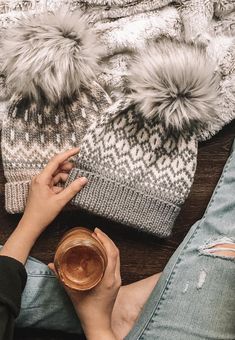 a woman sitting on the floor holding a cup of coffee and wearing a knitted hat with pom - poms