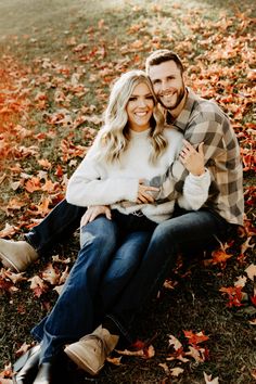 a man and woman sitting on the ground surrounded by fall leaves in an open field