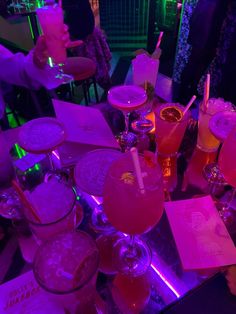 many different types of drinks on a table with purple lights in the background and people standing around