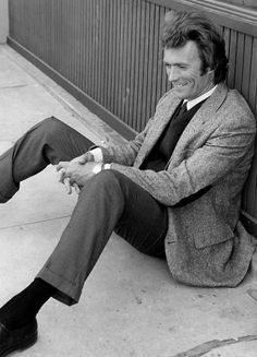 a man sitting on the sidewalk wearing a suit and tie with his legs crossed, smiling