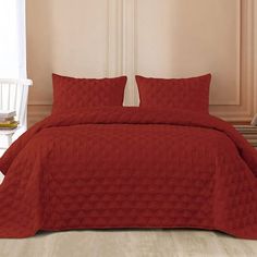 a bed with red bedspread and pillows in a room next to a white chair