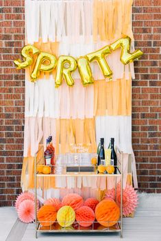 an orange and yellow party with balloons, streamers and drinks on a table in front of a brick wall