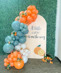 an arch made out of balloons and oranges on the grass with a sign that says a little cute is on the way