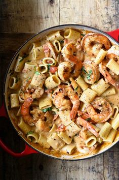 a red pot filled with pasta and shrimp on top of a wooden table next to a spoon