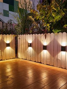 some lights are on the side of a wooden fence in front of plants and trees