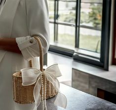 a woman holding a wicker basket with a white bow on the handle, standing in front of a window